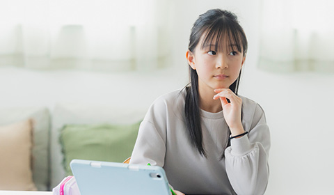 腕を机につき遠くを見て考える女の子 手前にはタブレットがある