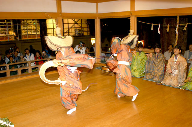 オレンジ色の衣装を着て、こきりこ楽器を持ち踊る2人の写真