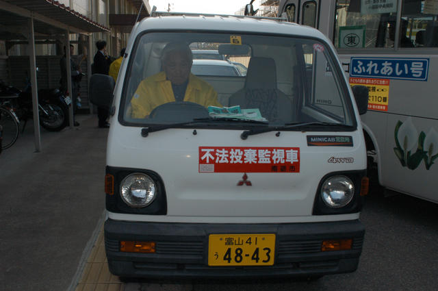 不法投棄監視車と書かれたステッカーが、黄色のジャンパーを着た男性が運転している軽トラックの全面に貼られている写真