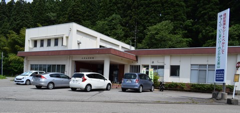停留場の近くに大きな看板があり、駐車場には車が4台、その奥に建つ南砺市太美山交流センターの入り口側から全体を撮影した写真