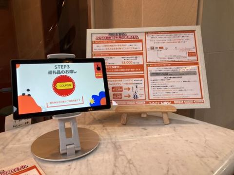 テーブルの上に額に入ったふるさと納税のチラシと、「STEP3返礼品のお渡し」と書かれた画面が表示されたタブレット端末が置かれている写真