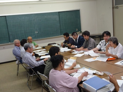 室内の中央に長机を寄せ、参加者が机の左側と右側に向かい合うように座り会議が行われている様子の写真