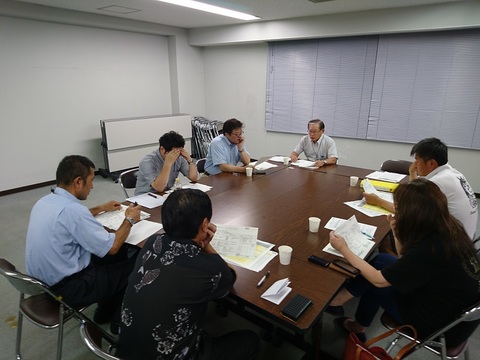7名の参加者が部屋の中央に寄せた長机の席に座り、それぞれ手元の資料を見ながら会議が行われている様子の写真