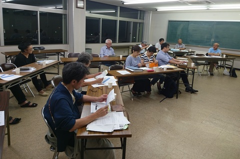 室内に扇状に設置された長机の席に参加者が座り、手元の資料を見ながら会議が行われている様子の写真
