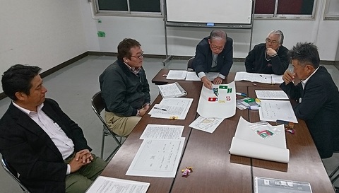同じテーブルを囲んで座っている5人の男性のうち1人の男性がテーブルに置かれている資料の紙をペンで指し、話し合いが行われている写真