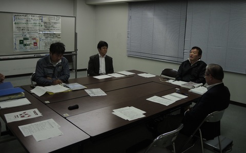 4人の男性参加者が寄せ合った6つの長机の席に座り、話し合いを行っている写真