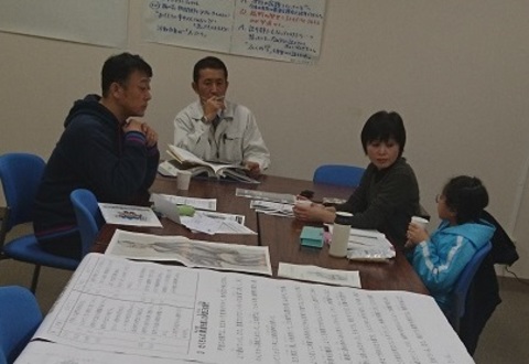 資料がたくさん置かれた机の席に女の子、女性、男性2名が座っている写真