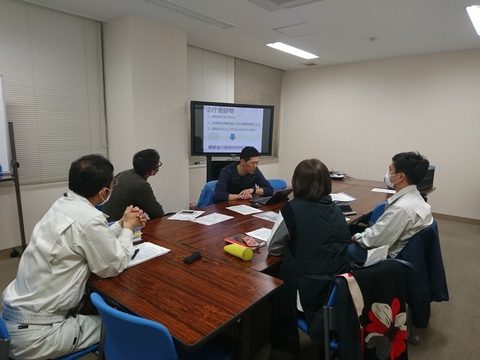 5名の参加者がグループになって席に座り、モニターに映し出された資料を見ながら会議が行われている写真