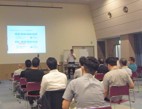 参加者達が設置された椅子に座り前方でマイクを持ち話をしている男性の話を聞いている様子の写真