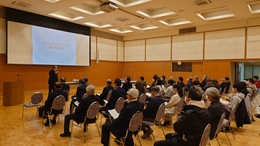 意見交換会で室内前方で話をしている男性、席に座り話を聞いている多くの参加者達を左後方から撮影した写真