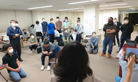 立っている参加者、床に座っている参加者、パイプ椅子に座っている参加者が手前にいる女性の話を聞いている写真