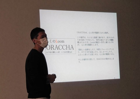 文字が映されたスクリーンが投影された壁の前に立ち話をする男性の写真