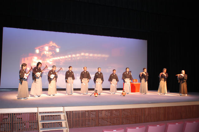 和装姿の男性たちが舞台の上で三味線や笛を演奏している定期公演の様子を写した写真