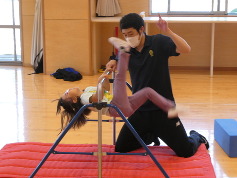 ちいさな子供が鉄棒を回ろうと足を振り上げ補助の男性が教えているなんと元気っ子教室！の写真