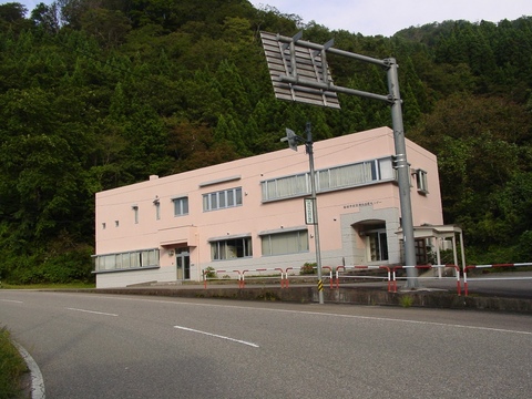 左側にカーブした道路と木々が鬱蒼と茂った林の間に立つ、薄いピンク色の外壁の診療所の建物外観の写真