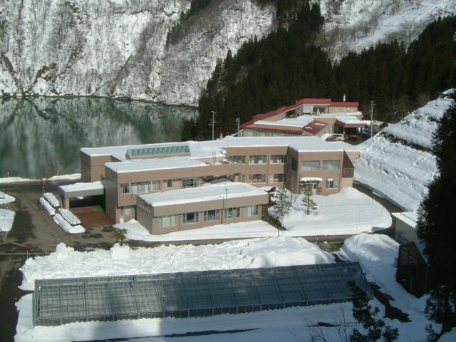 雪が積もった山の麓の池の手前に立つ、ベージュ色の外壁のつつじ荘の建物外観の写真
