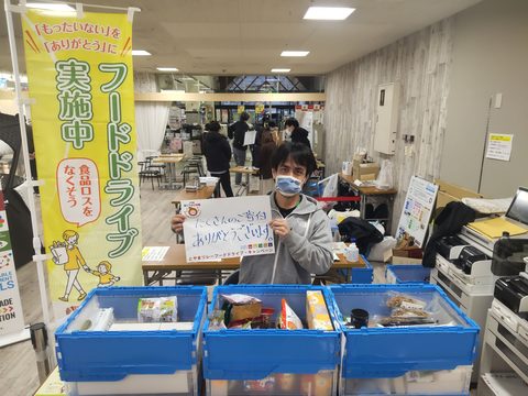 「フードドライブ実施中」と書かれたのぼり旗が立っている横に食料品が入った3つの容器が並べられ、その後ろで男性が「たくさんのご寄付ありがとうございます。」と書かれた紙を持っている写真
