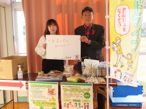 長机の上にインスタントフード等が置かれ、机の後ろに立っている女性と年配の男性が「食品ロスの助け合い!!」と書かれた紙を一緒に持っている写真