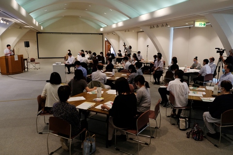 天井が丸みを帯びている広い室内で、グループごとに分かれて席についている参加者達が、左側に設置された演台で話をしている男性講師の方の話を聞いている写真