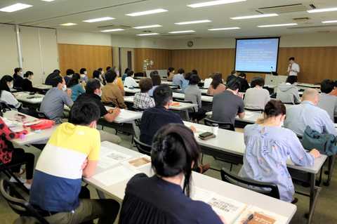 設置された長机の席に参加者が2名ずつ座り、前方に映し出されたスクリーンを見ながらスクリーン横に立っている男性の話を聞いている様子の写真