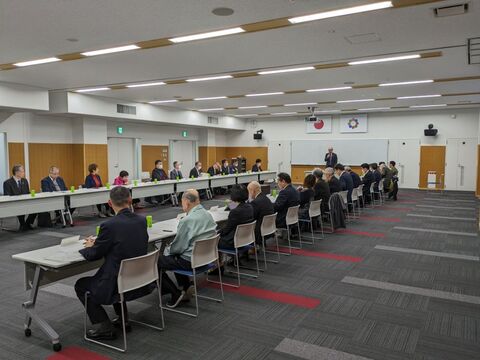 前方にスーツを着た男性が立って話しているのを、コの字型に設置されたテーブルと椅子に座り話を聞いている様子の写真